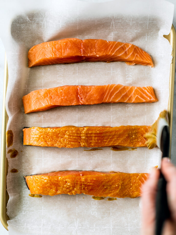 Raw filets of salmon on a parchment lined baking sheet being brushed with miso glaze.