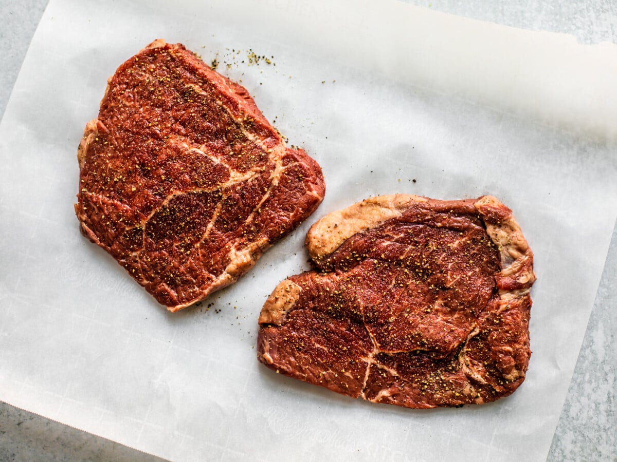 Two raw tenderized and seasoned steaks on parchment paper.