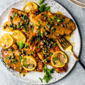Platter of chicken piccata smothered in sauce, capers, lemon slices, and fresh parsley.