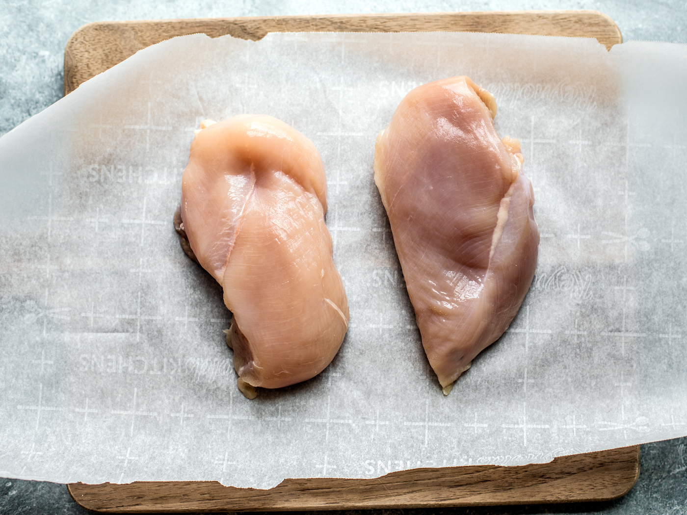 Two raw chicken breasts on parchment-lined cutting board.