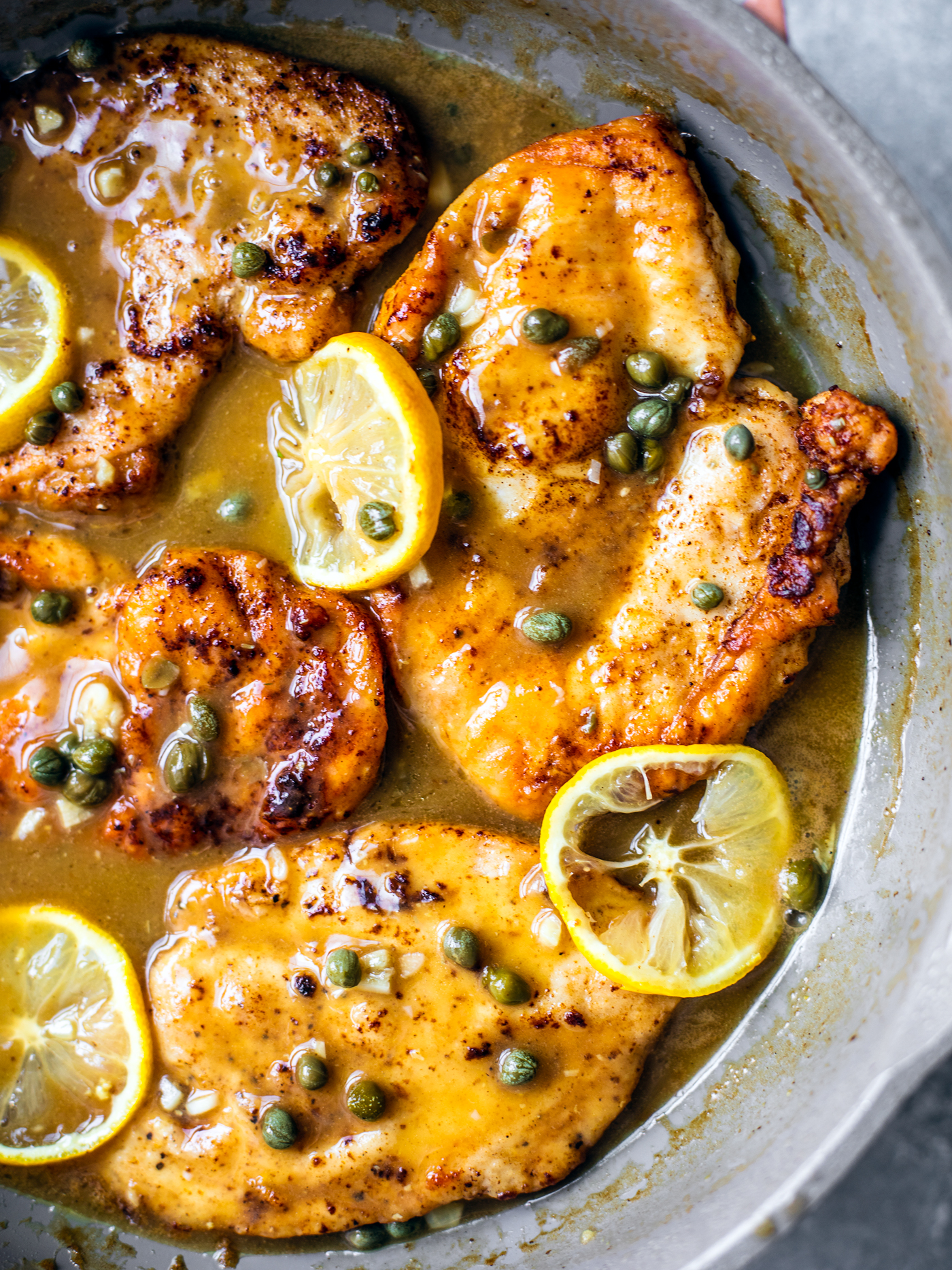 Close up of golden chicken cutlets in skillet with sauce, capers, and lemon slices.