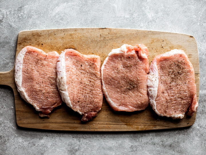 Four tenderized pork chops lined up on a cutting board, seasoned.