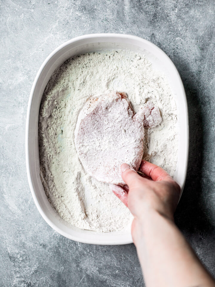 Hand taking floured pork chop out of dish of flour.
