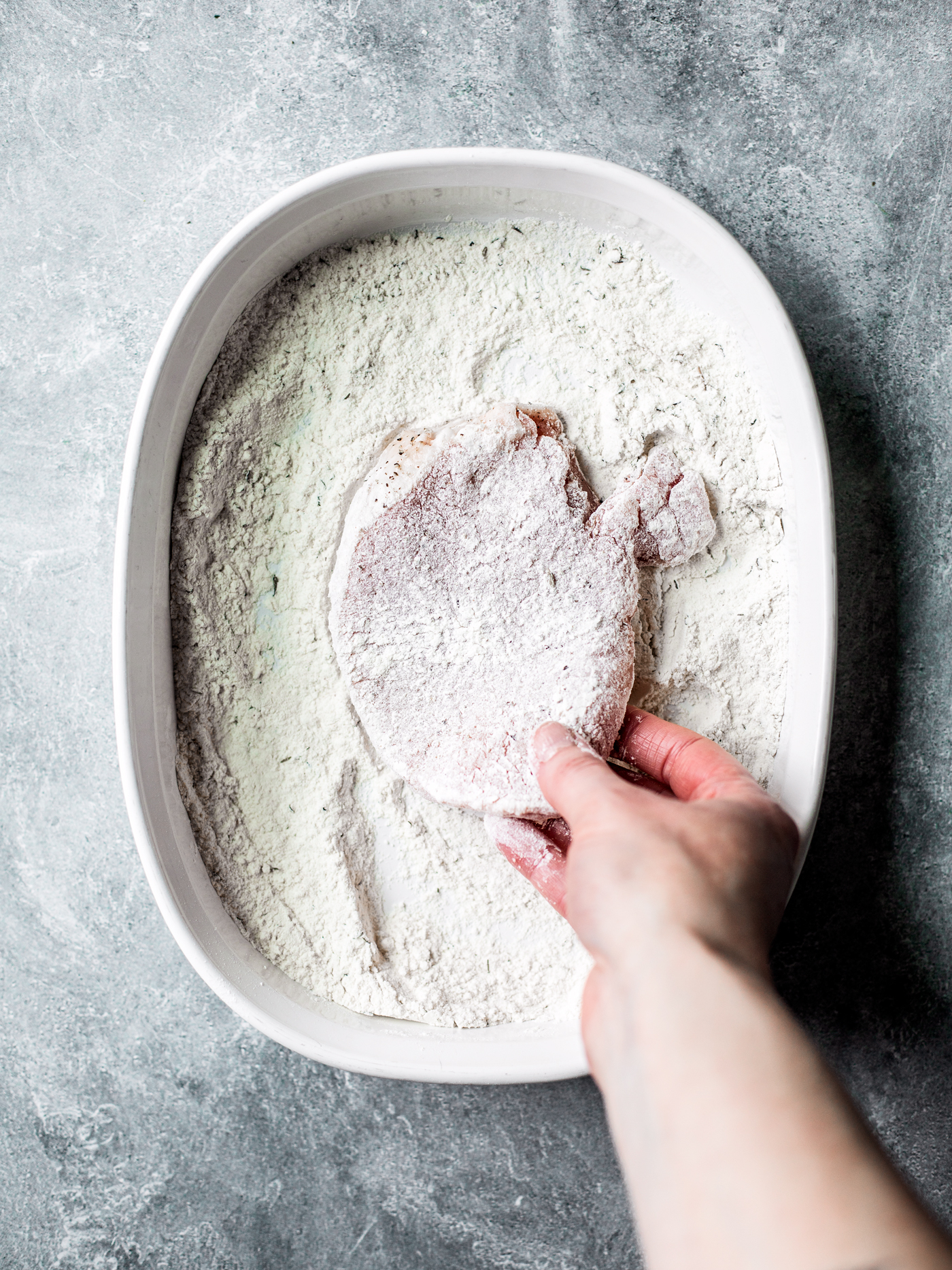 Hand taking floured pork chop out of dish of flour.