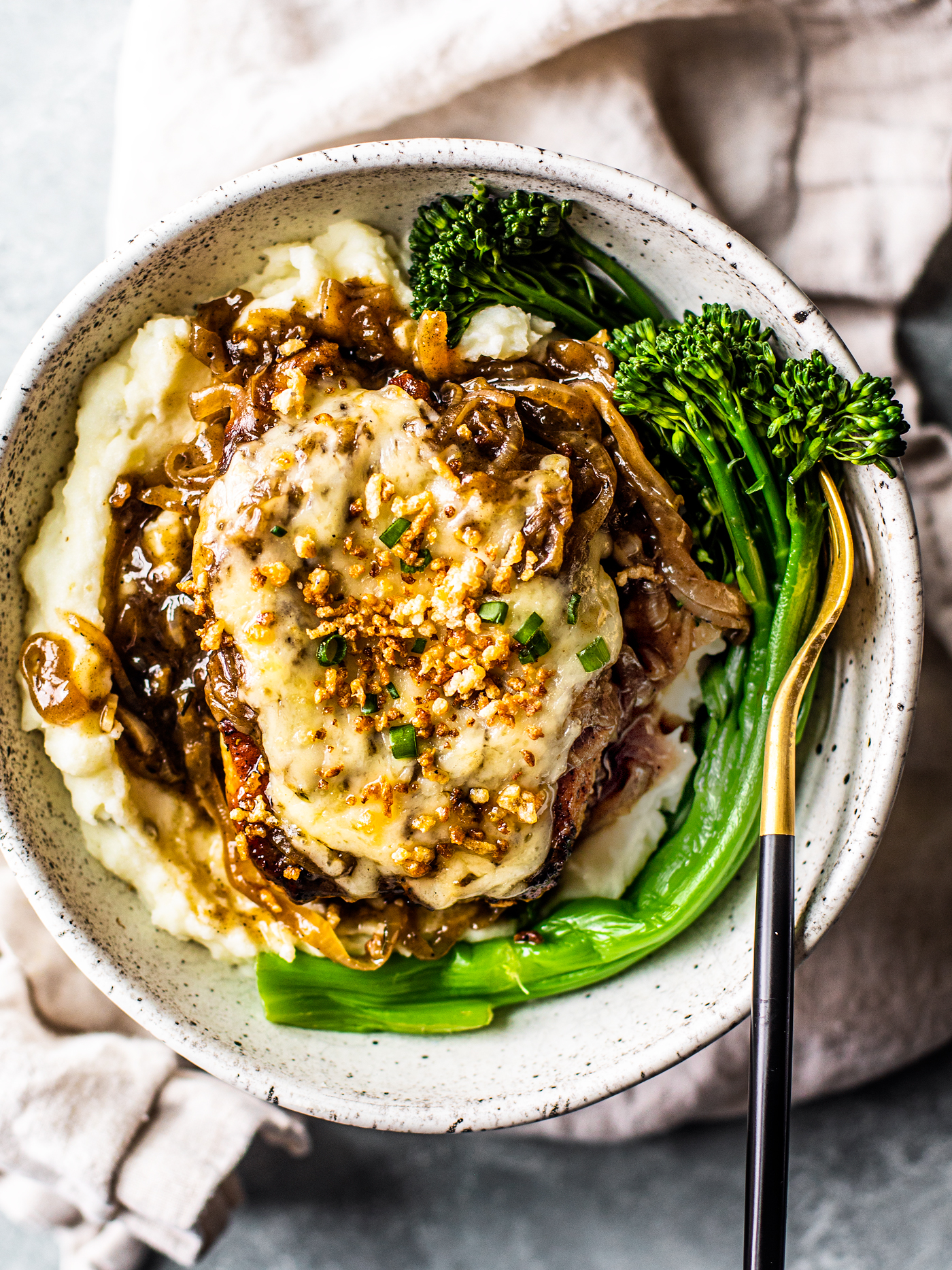 Cheese-topped pork chop with gravy on top of mashed potatoes in a bowl with broccolini.