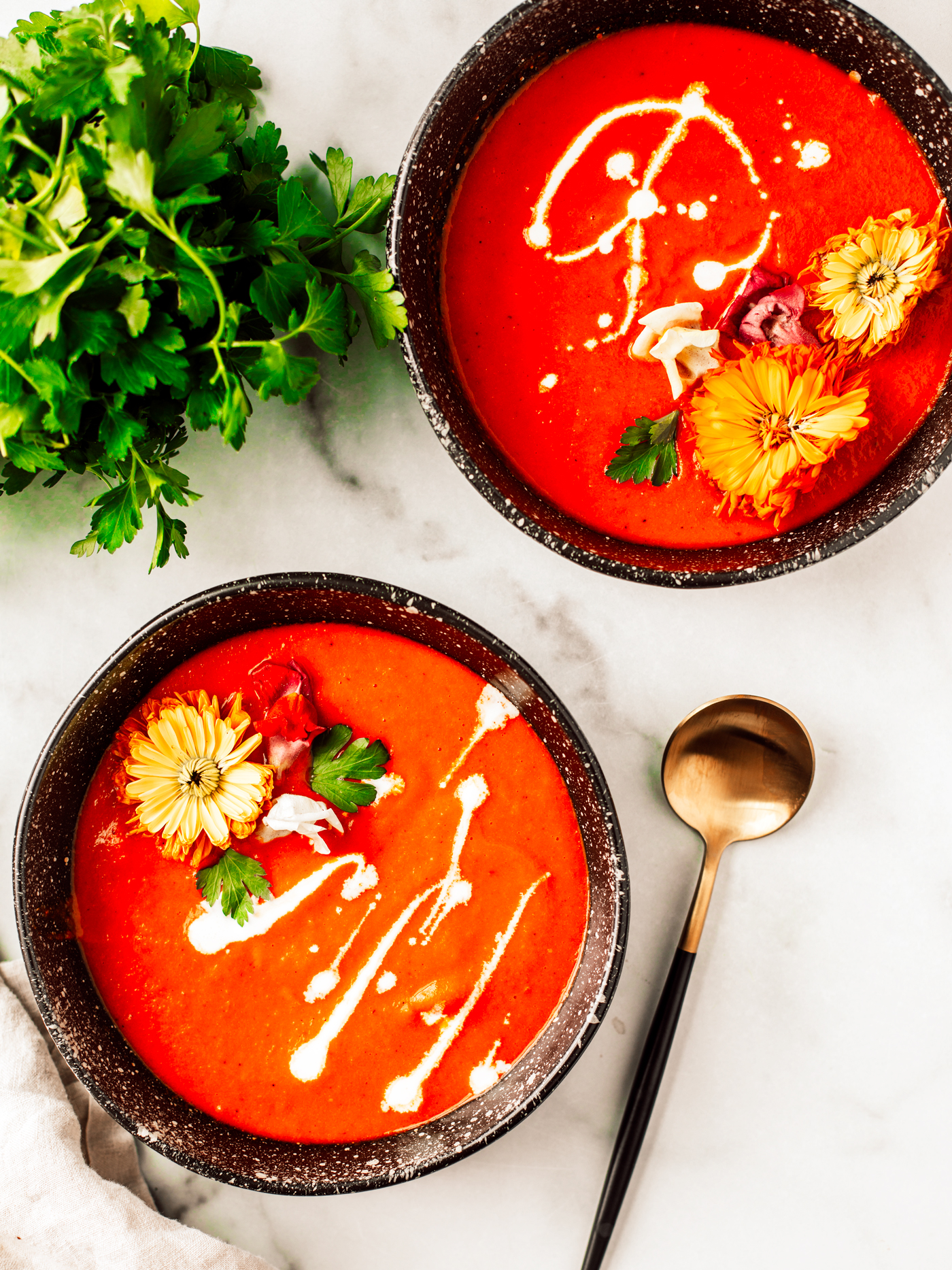 Two bowls of roasted red pepper soup garnished with drizzled cream and edible flowers.