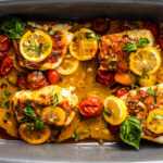 Gray baking dish with four cod fillets in tomato herb butter topped with fresh herbs and lemon slices.