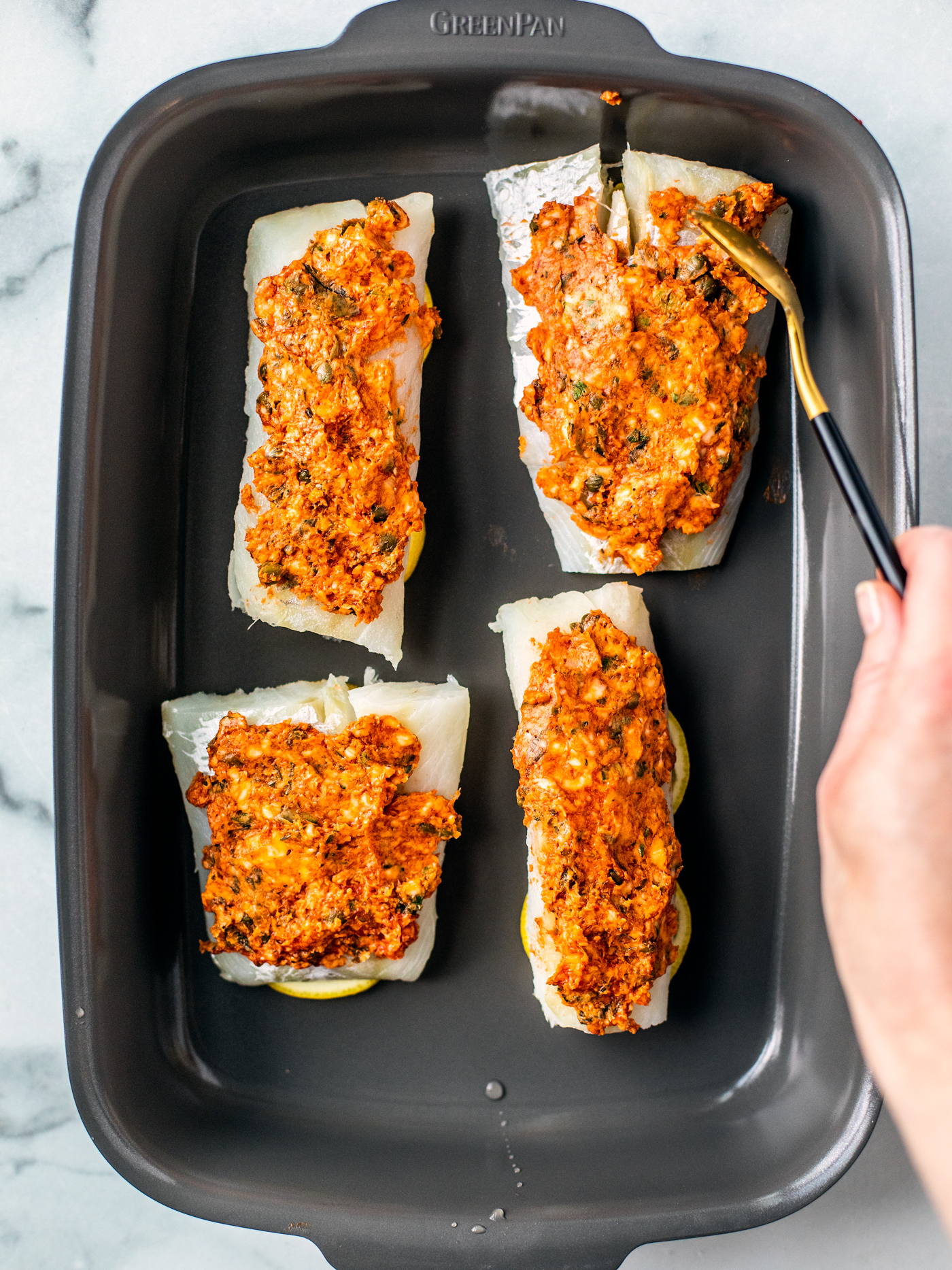 Baking dish with four cod fillets being topped with compound tomato butter.