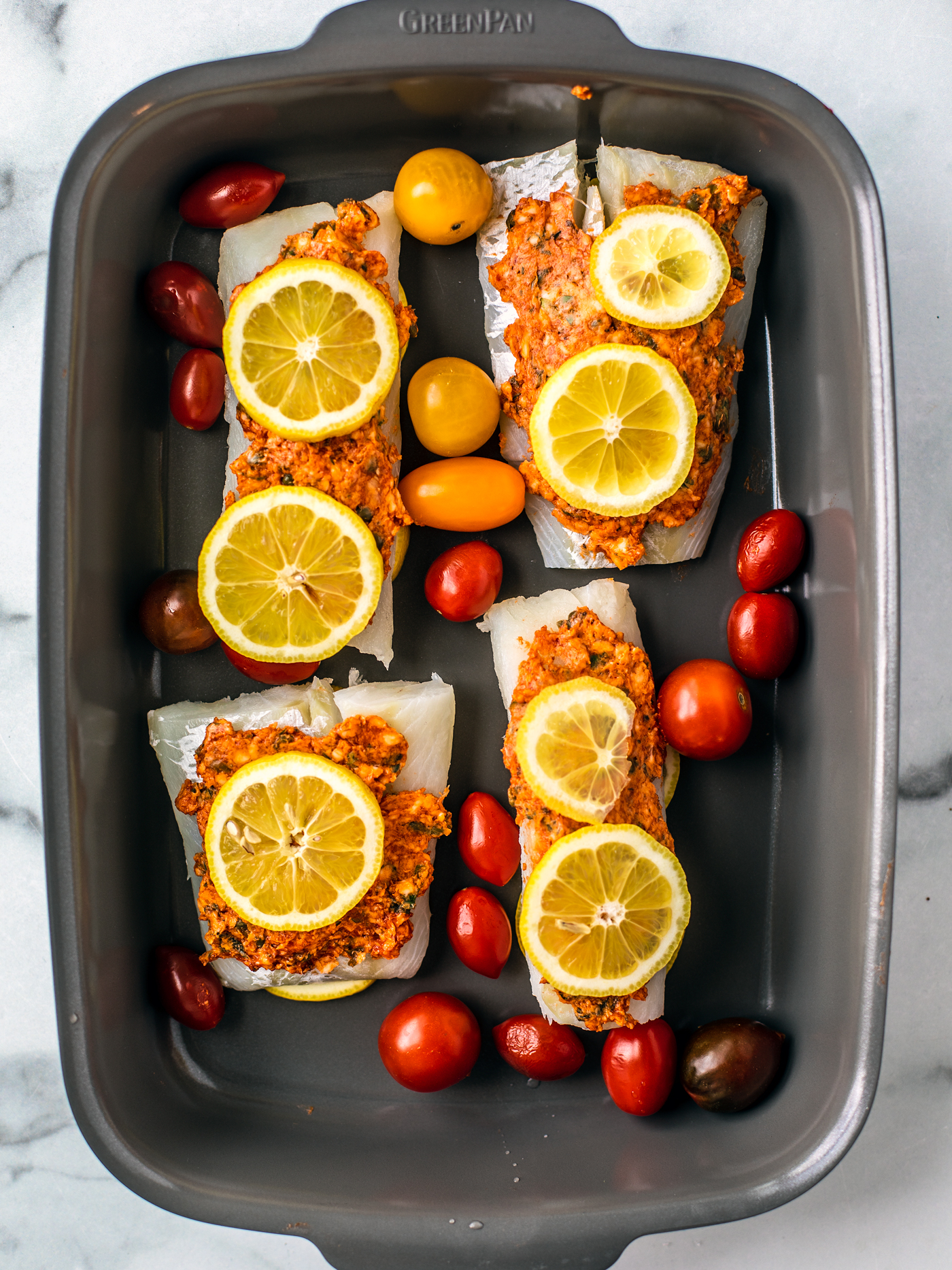 Baking dish with four cod fillets topped with compound butter and lemons with grape tomatoes on the side.