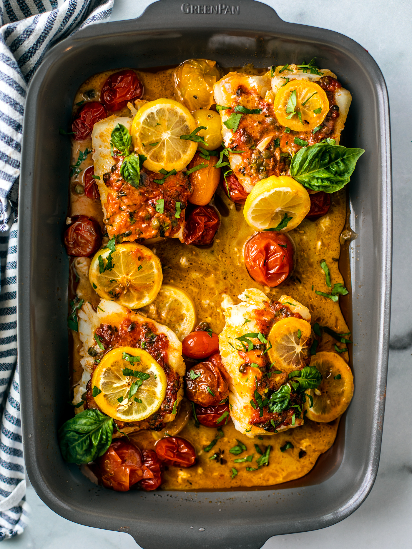Gray baking dish with four cod fillets in tomato herb butter topped with fresh herbs and lemon slices.