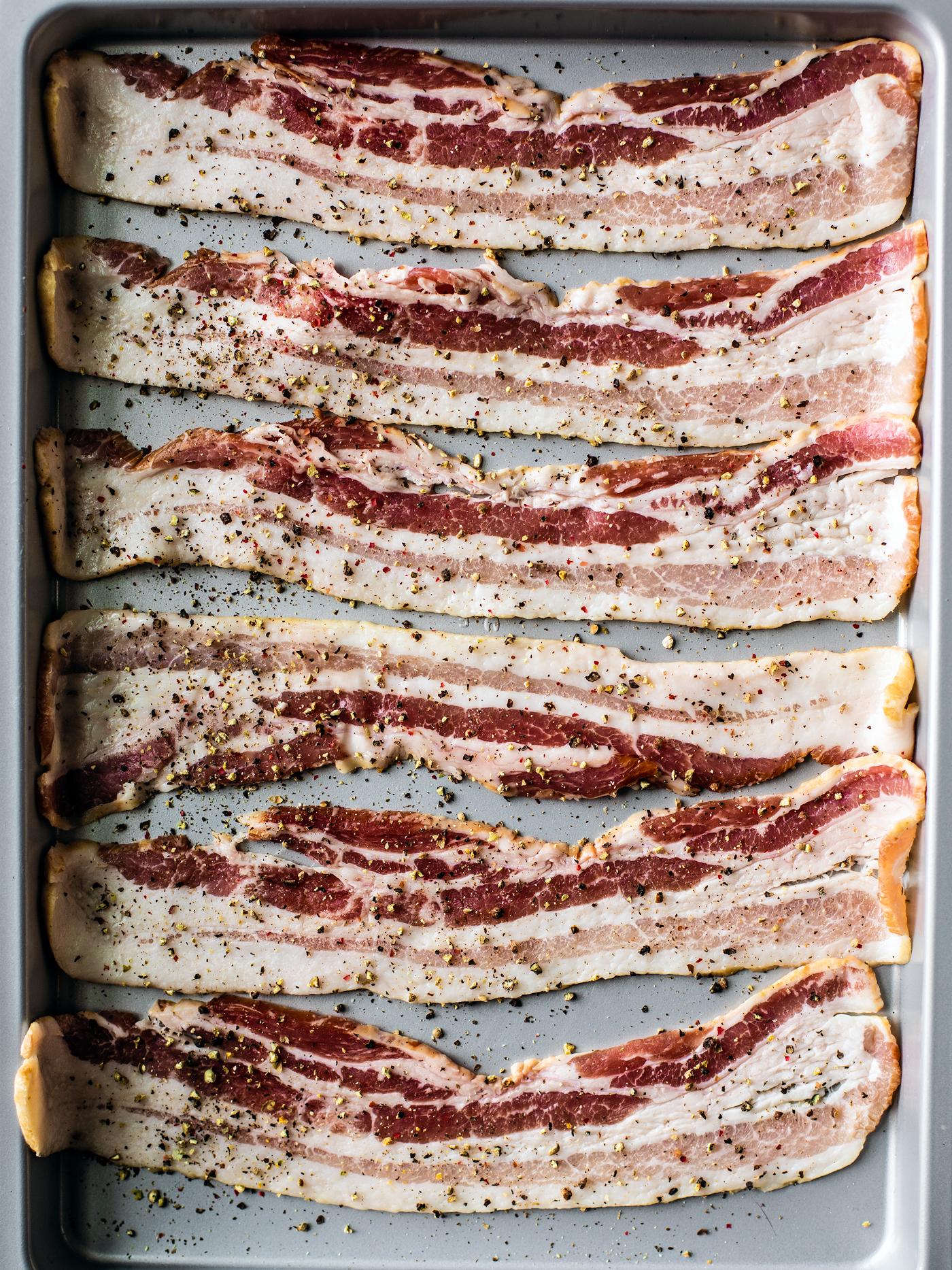 Baking sheet of uncooked bacon with cracked black pepper.