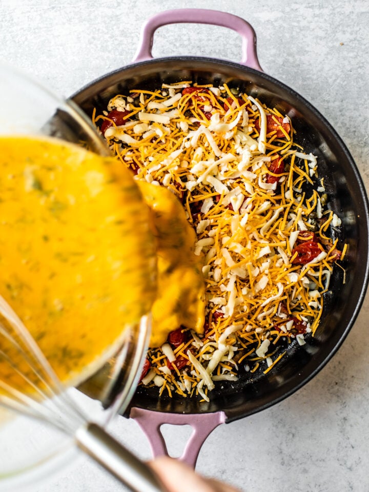 Egg mixture being poured over veggies and cheese in cast iron skillet.
