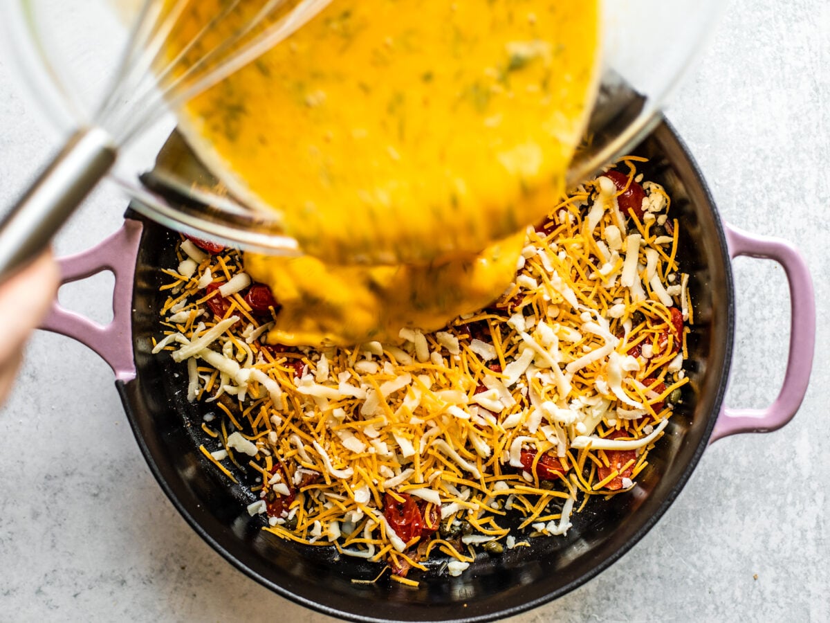 Egg mixture being poured over veggies and cheese in cast iron skillet.