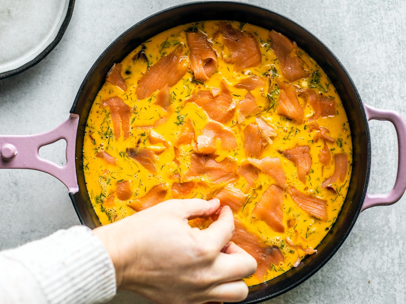 Hand placing piece of smoked salmon into egg mixture in skillet.