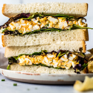 Egg salad sandwich halves with mixed greens stacked on a plate with a side of chips.