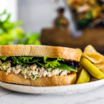 SIde shot of tuna sandwich with greens on plate with pickles and chips.