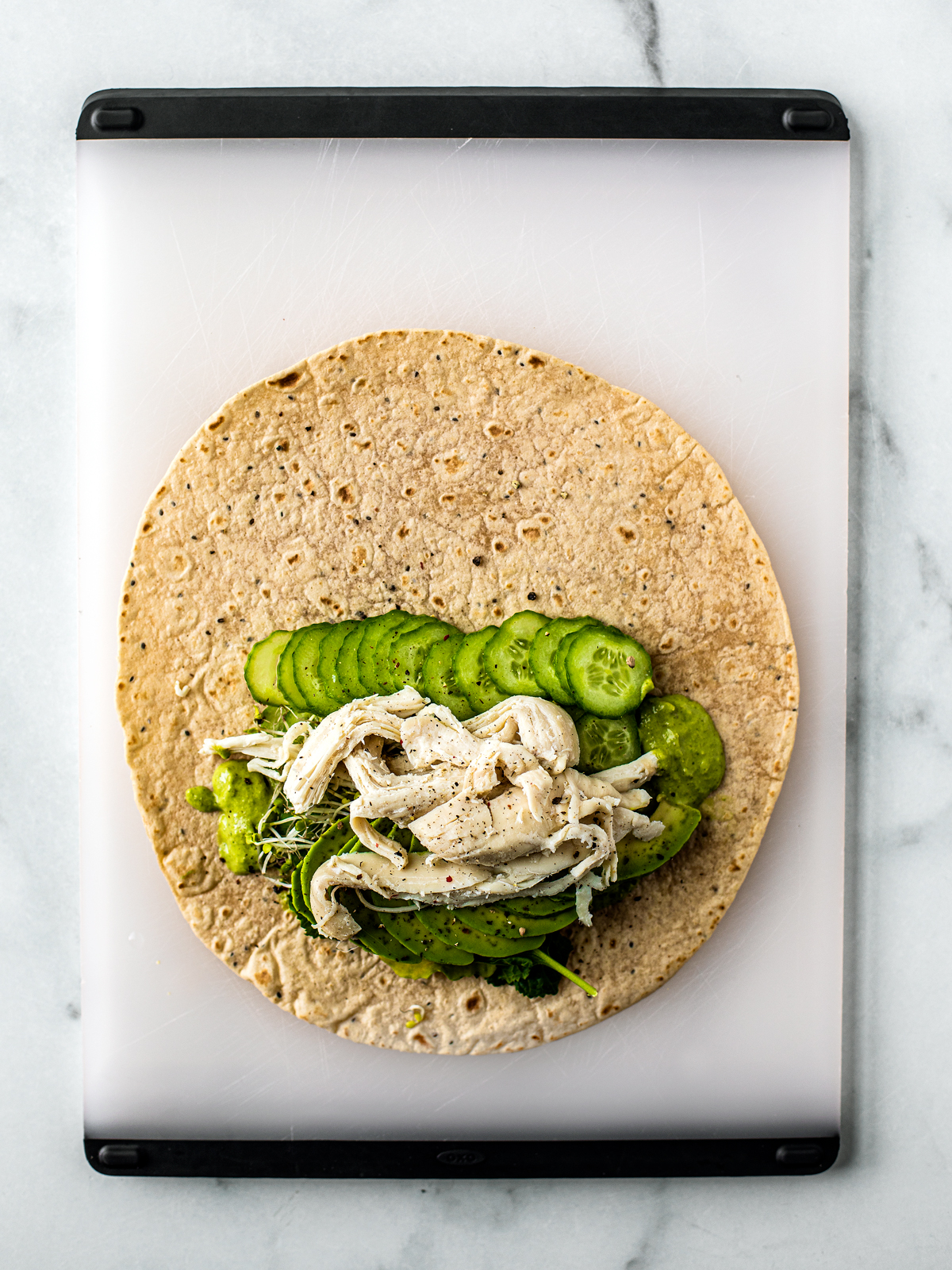 Wrap on cutting board with chicken, cucumbers, greens, avocado, and sprouts on it.