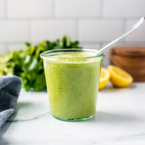 A jar of bright green dressing with lemons and parsley in the background in front of a white-tiled wall.