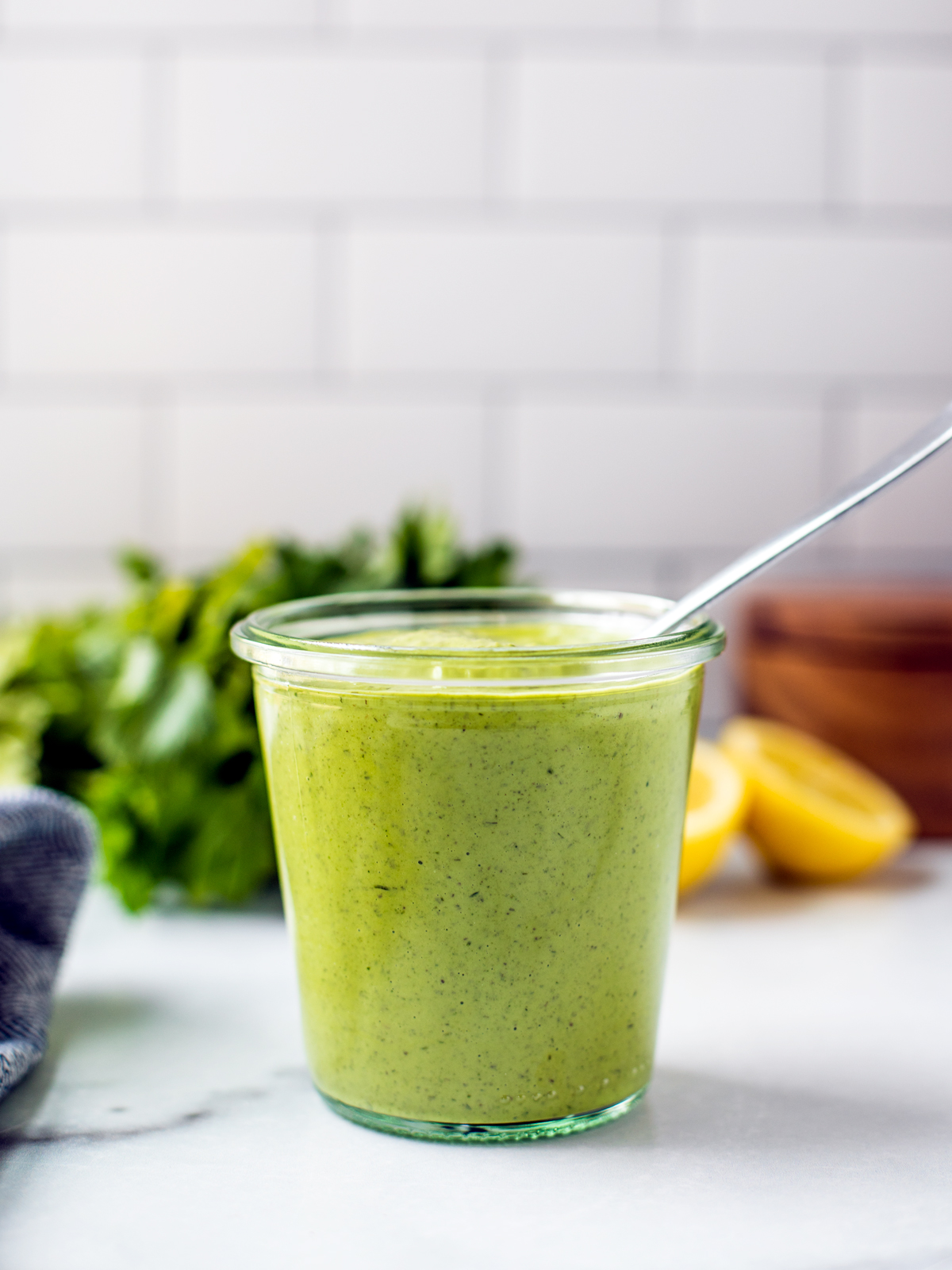 A jar of bright green dressing with lemons and parsley in the background in front of a white-tiled wall.