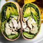 Close up of green goddess wrap on a white plate with a side of chips and cucumber spears.