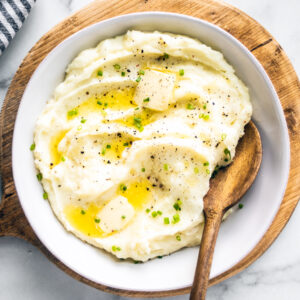 White serving bowl with creamy butter mashed potatoes with a wooden spoon set into it.