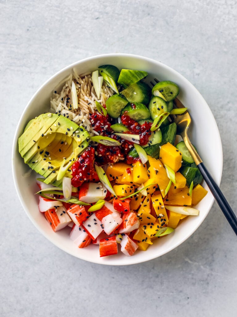 White bowl of rice, avocado, cucumbers, mango, and crab.