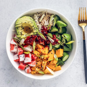 White bowl of rice, avocado, cucumbers, mango, and crab with a fork on the side.