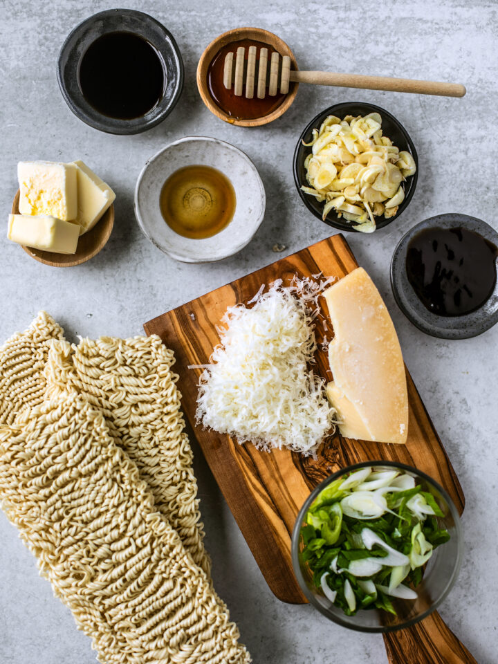 Dry Chinese noodles on a countertop next to small bowls of ingredients and a small cutting board with grated parmesan cheese.