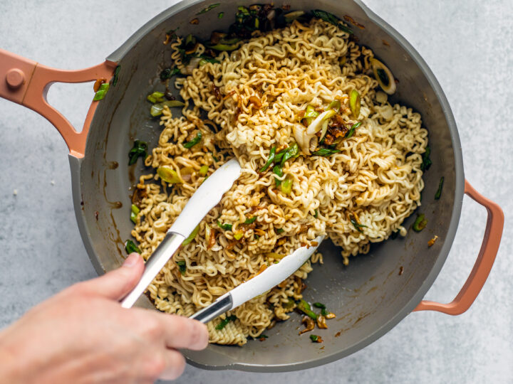 Hand tossing noodles in skillet with tongs.