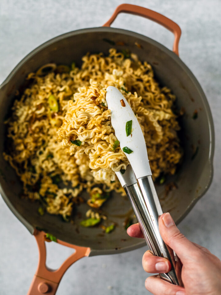 Hand tossing noodles in skillet with tongs.
