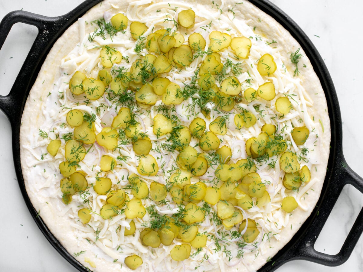 Uncooked pizza with cheese and pickle slices.