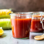 Jar of homemade BBQ sauce with a spoon in it with lemons and corn on the cob in the background.