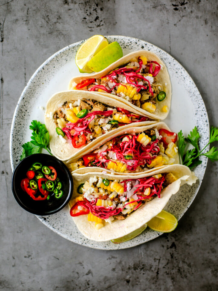 Plate of pulled pork tacos with colorful toppings and a pinch bowl of hot pepper slices.