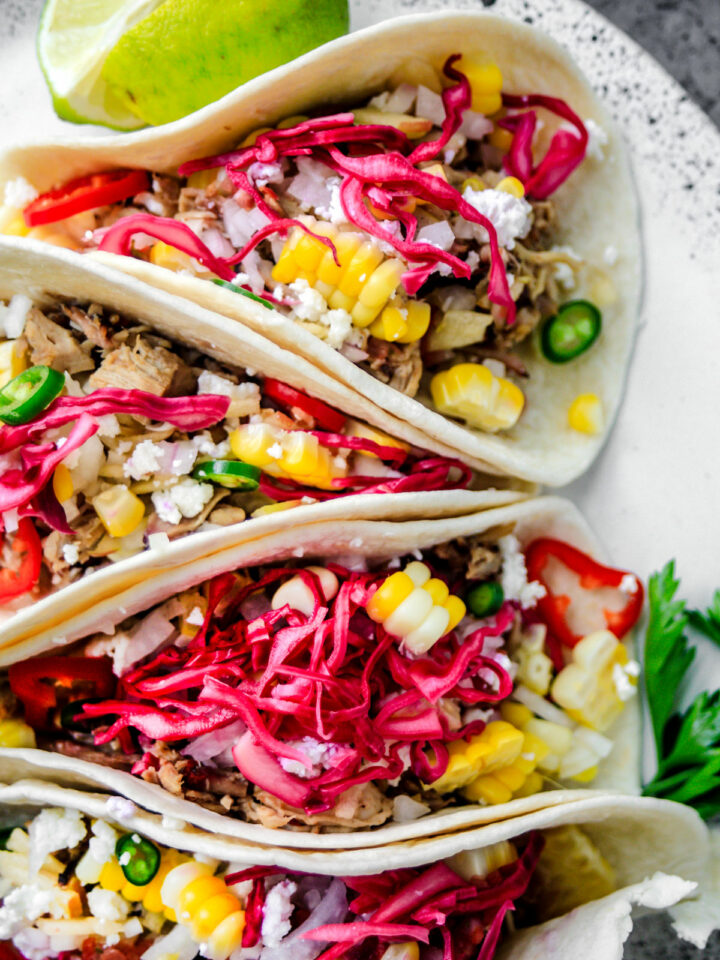 Close up of pulled pork tacos topped with red cabbage, corn, and hot peppers.