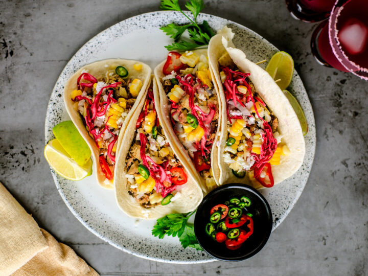 Plate of pulled pork tacos with colorful toppings and a pinch bowl of hot pepper slices.