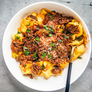 White bowl of pappardelle pasta tossed in pork ragu, garnished with parsley.