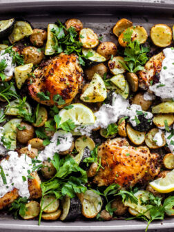 Sheet pan covered in roasted chicken thighs, zucchini, potatoes, with a drizzle of yogurt sauce and fresh herbs.