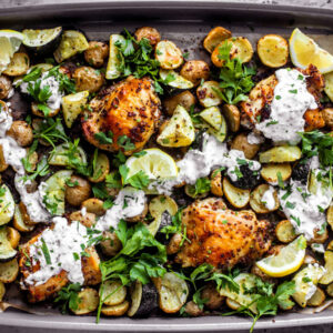 Sheet pan covered in roasted chicken thighs, zucchini, potatoes, with a drizzle of yogurt sauce and fresh herbs.