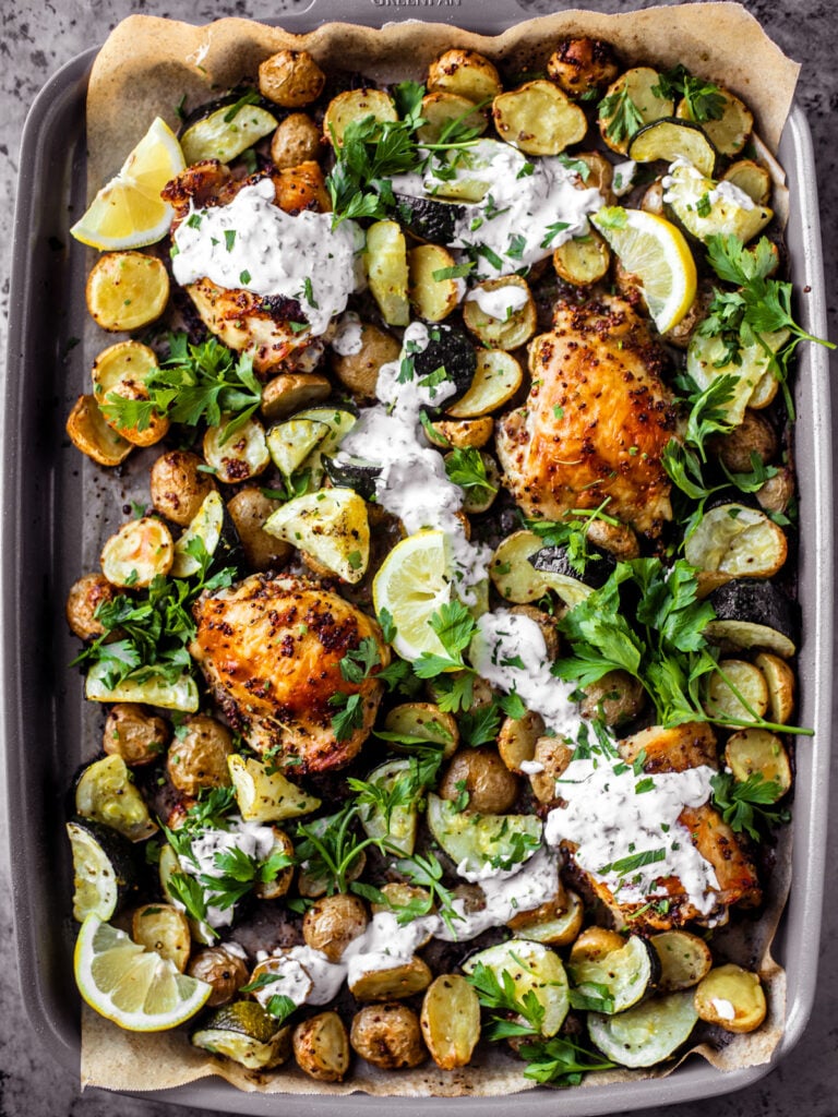 Sheet pan covered in roasted chicken thighs, zucchini, potatoes, with a drizzle of yogurt sauce and fresh herbs.
