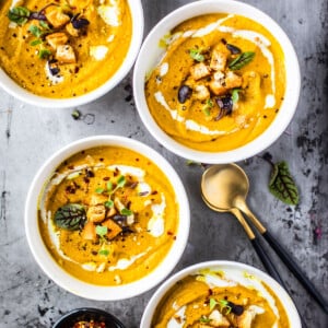 Four bowls of pumpkin cauliflower soup garnished with croutons, greens, and a cream drizzle.