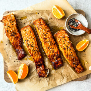 Four fillets of salmon with orange honey glaze on a serving board with orange wedges.