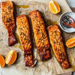 Four fillets of salmon with orange honey glaze on a serving board with orange wedges.