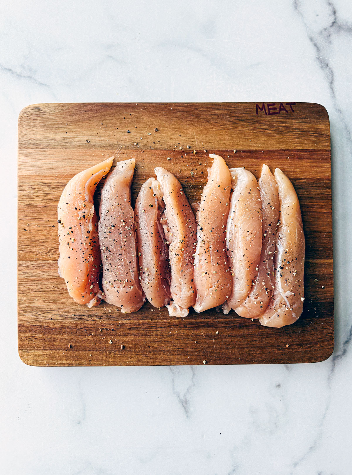 Wooden cutting board with uncooked chicken tenderloins on it.