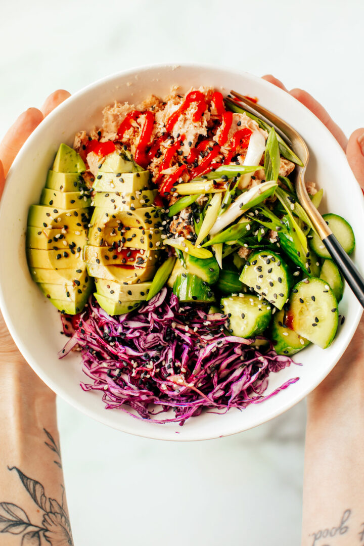 Hands holding a big tuna bowl with canned tuna, avocado, red cabbage, and cucumbers.