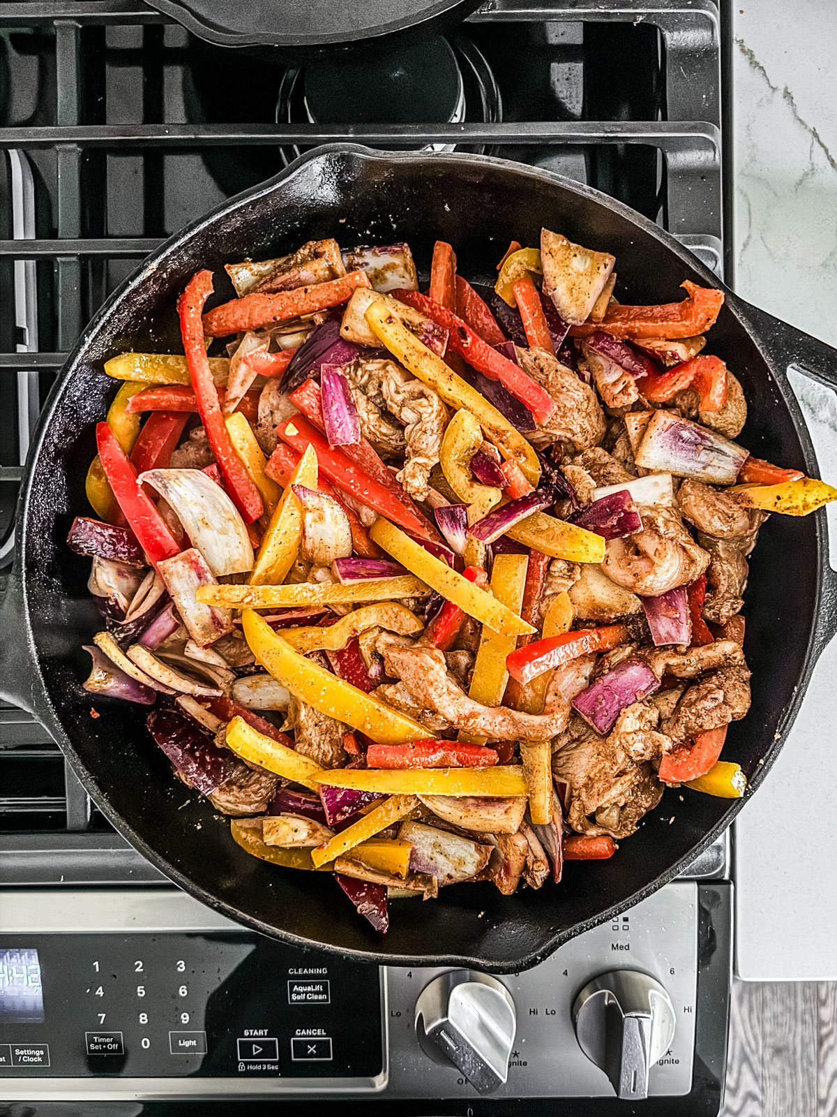 Skillet on stovetop with chicken strips, peppers, and onions.