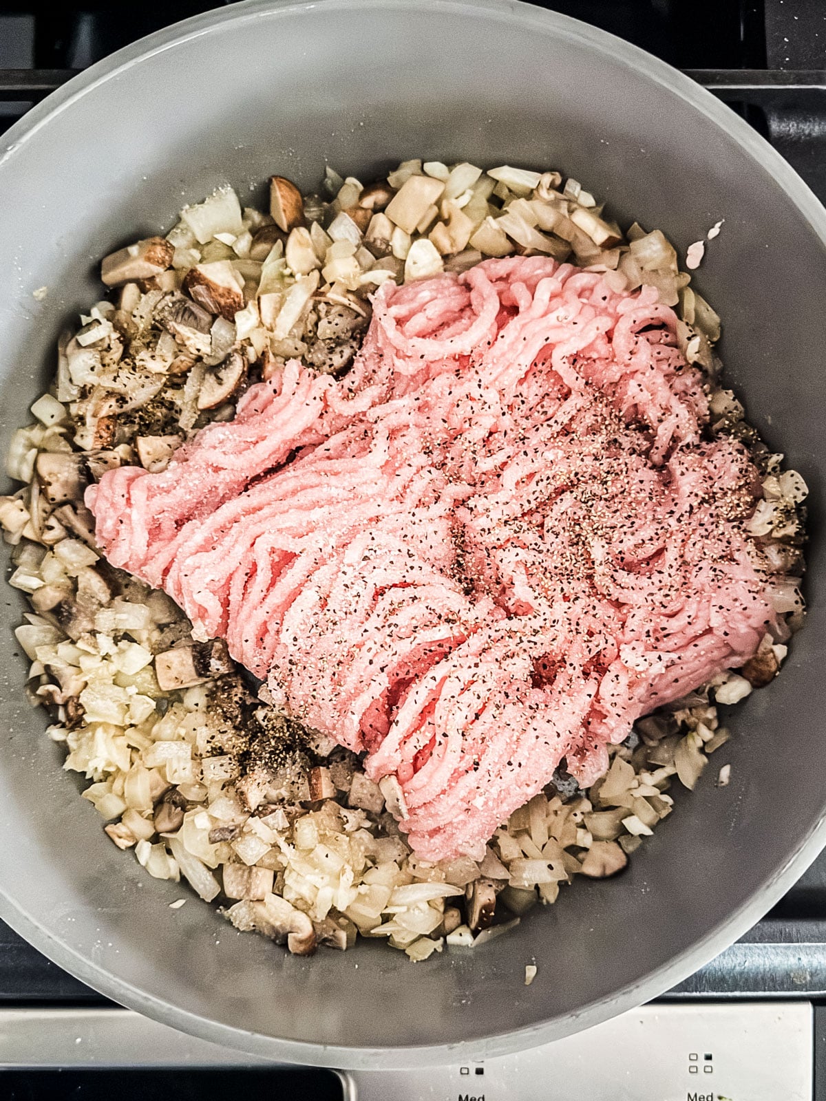 Uncooked ground turkey in pan with cooked onions and mushrooms.