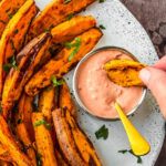 Air fryer sweet potatoes with one being dipped into spicy dipping sauce.