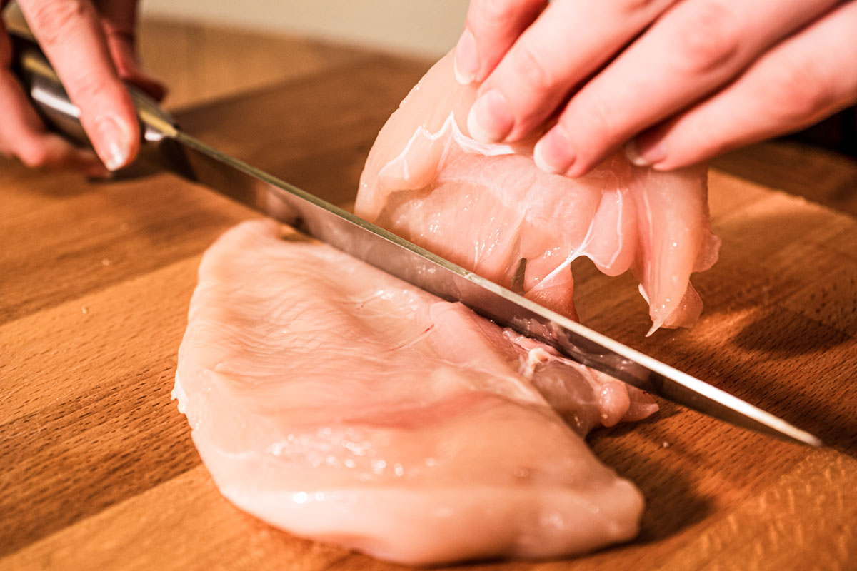 A chicken breast being cut into a cutlet on a cutting board.
