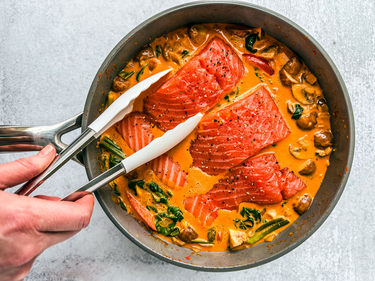 Salmon fillets being nestled into the curry sauce with tongs.