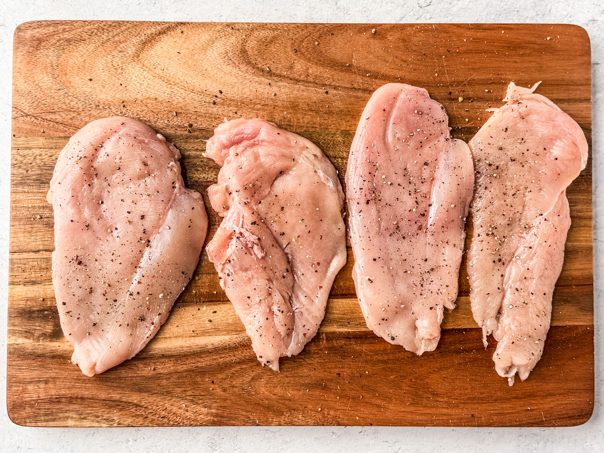 Chicken cutlets seasoned with salt and pepper laid out on a cutting board.